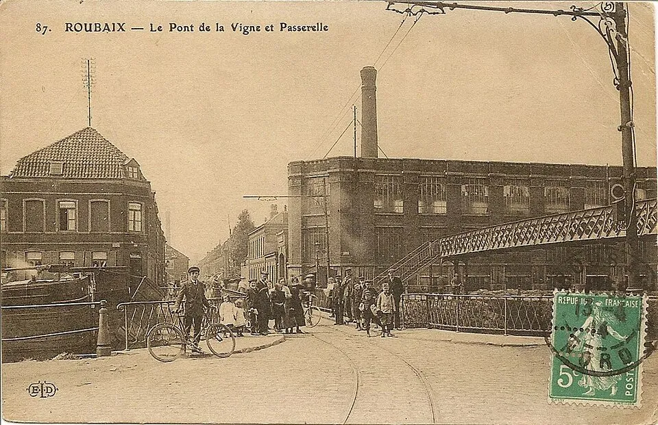 Carte postale sépia : un groupe d’adultes et d’enfants, dont un cycliste, au pied d’une passerelle métallique, devant une usine à haute cheminée