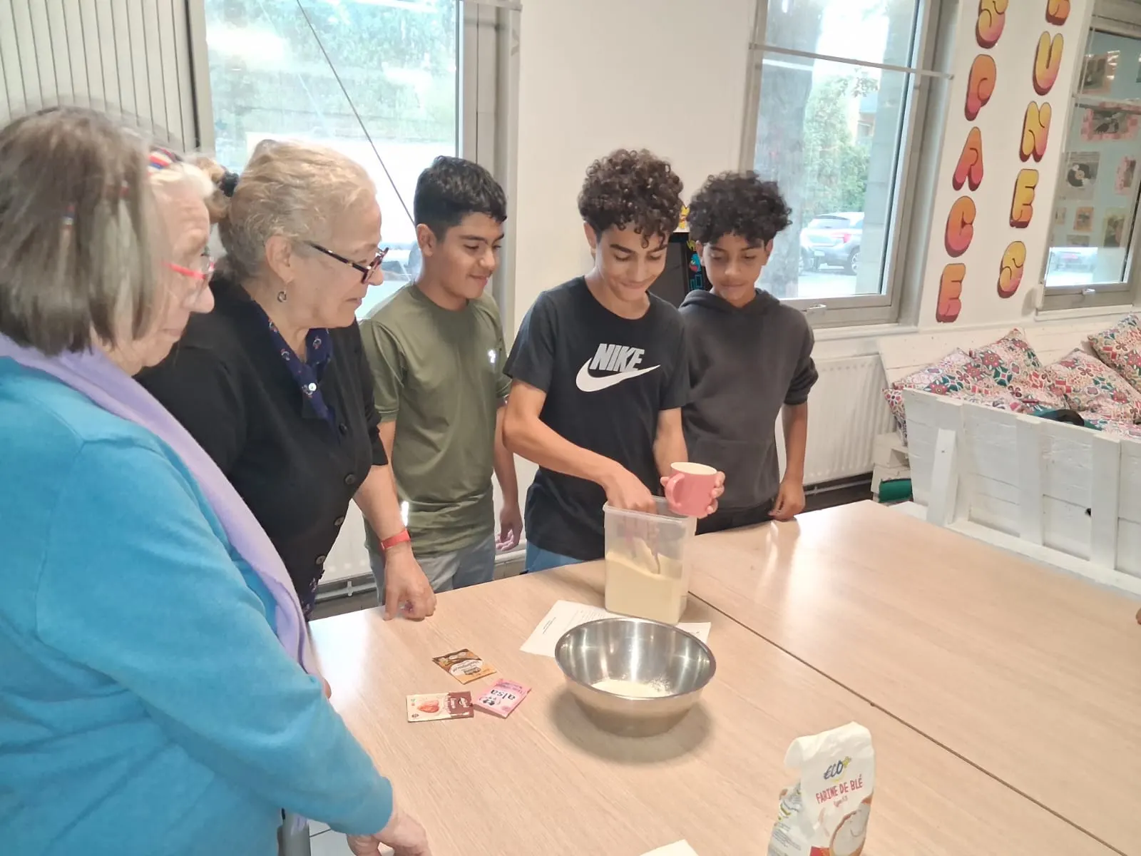 Un adolescent mesure la farine avec une tasse, sous le regard de deux seniors et de ses camarades