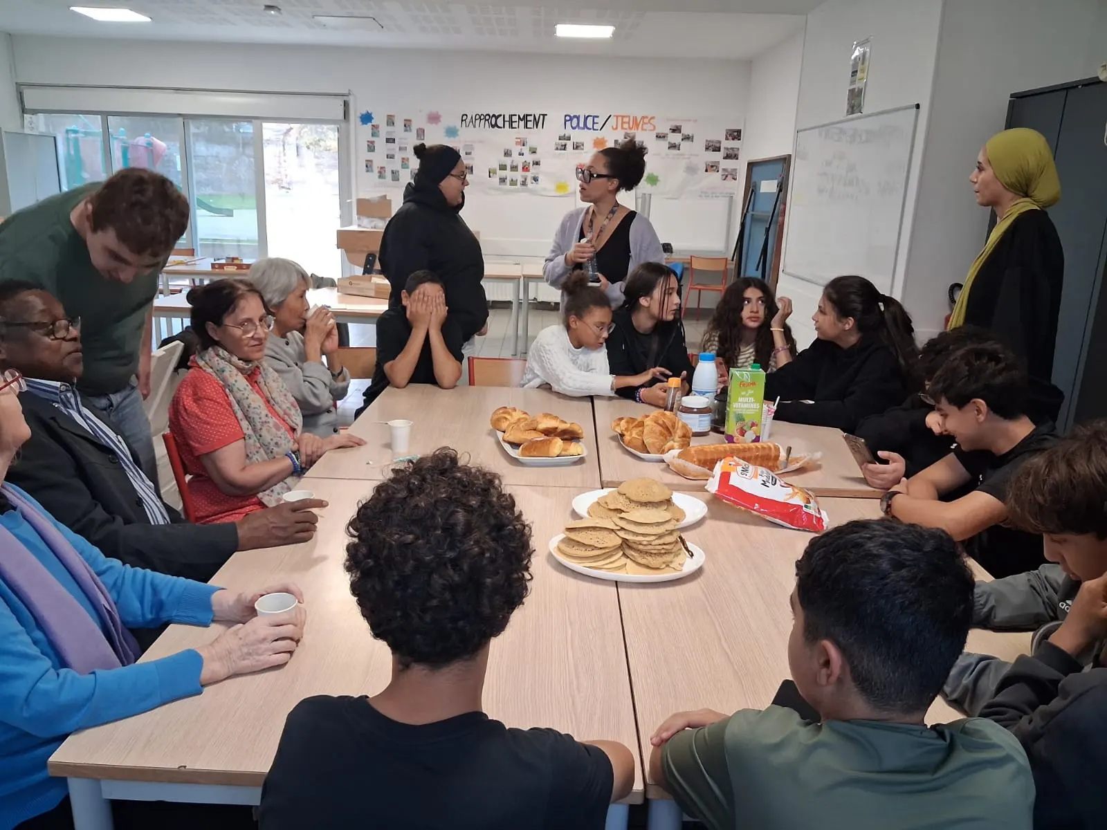 Seniors, adolescents et animatrices réunis autour d’une grande table garnie de crêpes et de viennoiseries