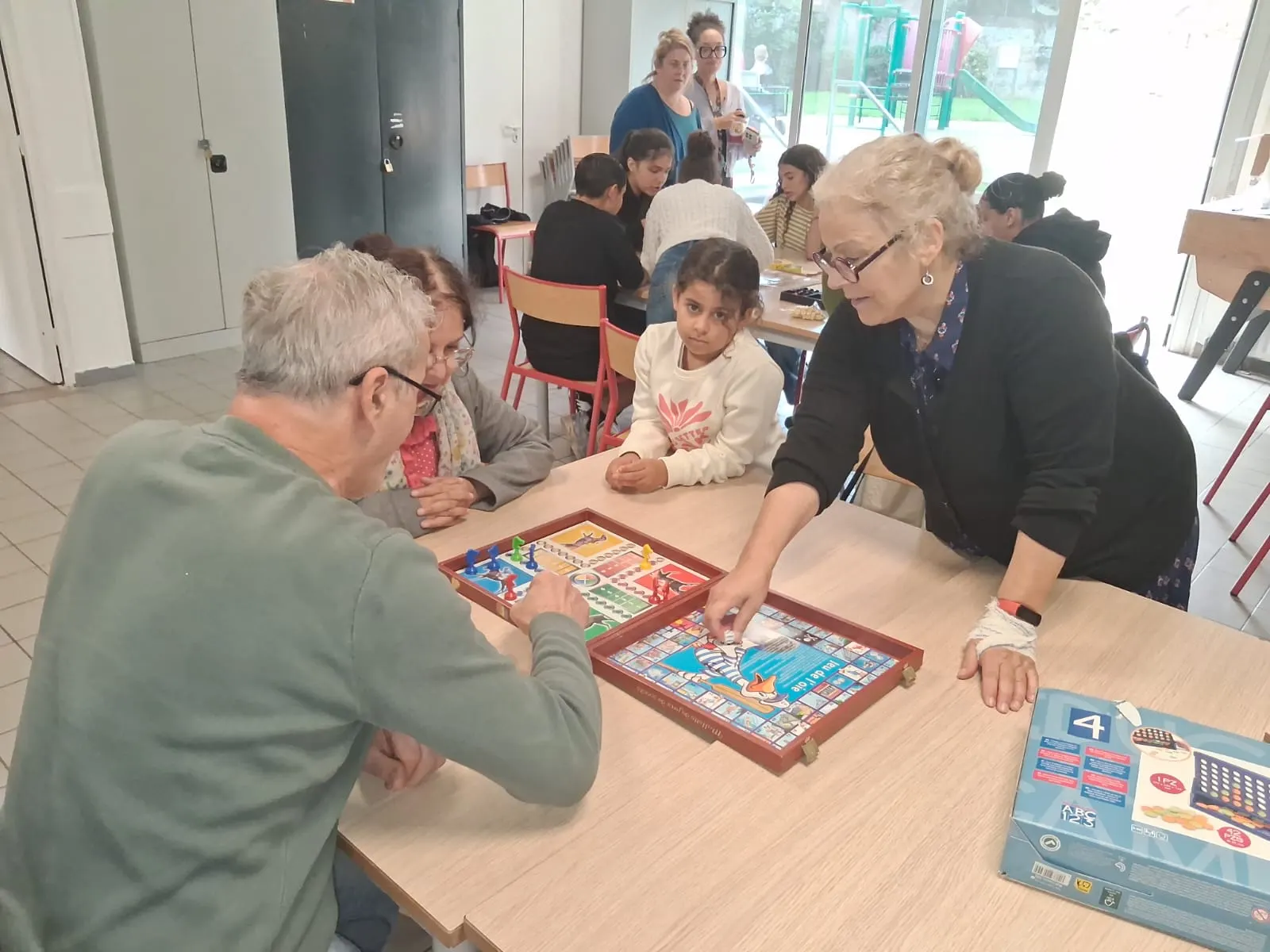 Une animatrice montre une case du jeu de l’oie à deux seniors et à une petite fille
