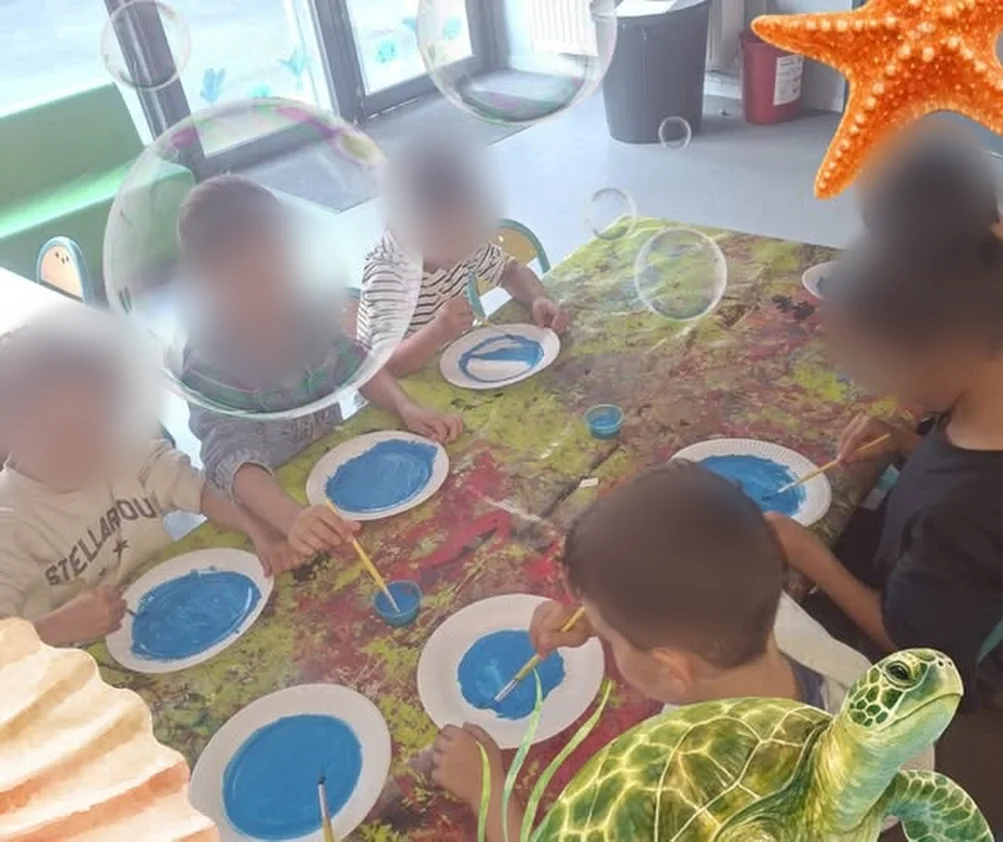 Des enfants peignent des assiettes en bleu sur une table couverte de peinture (visages floutés)