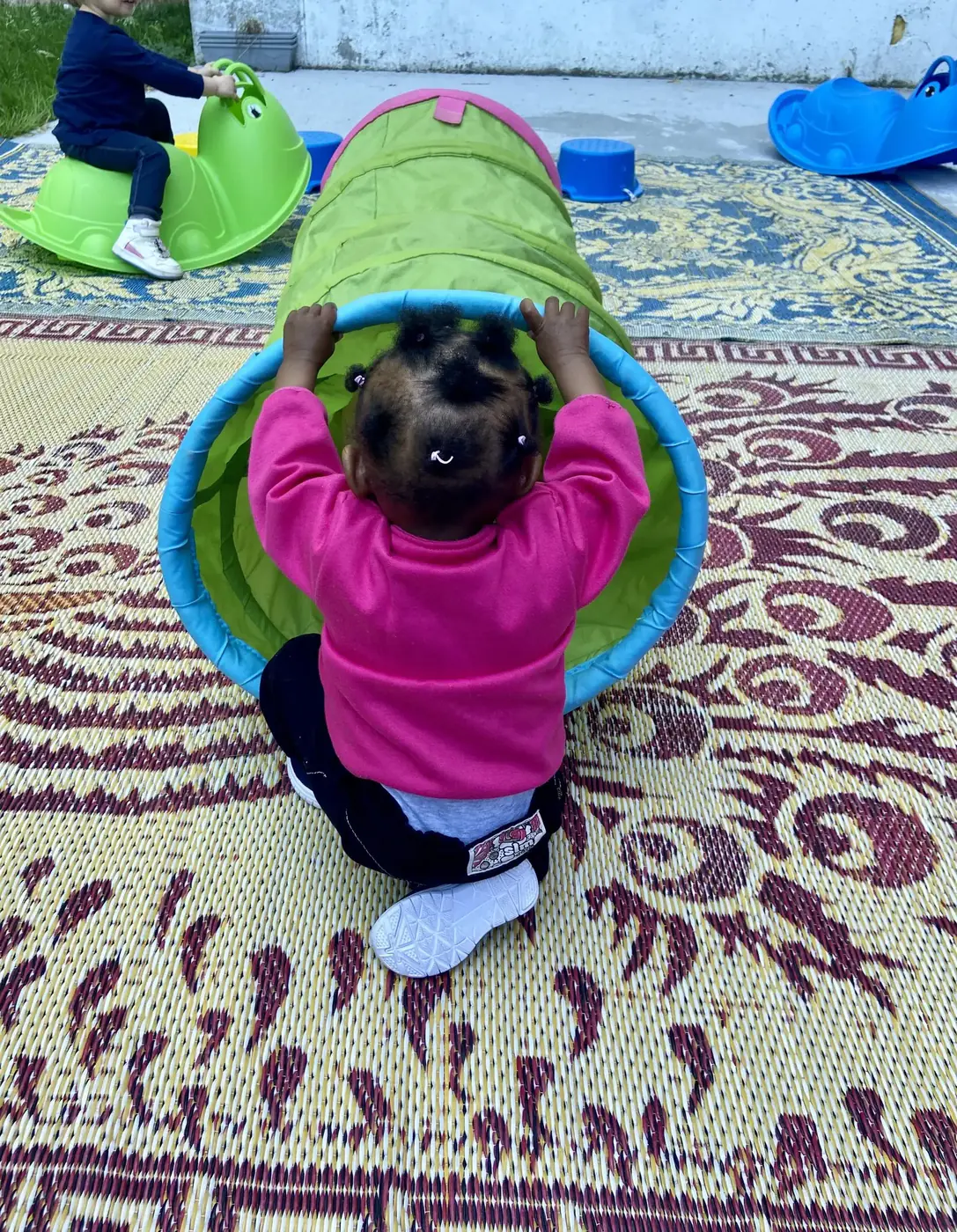 Un enfant, vu de dos, s’apprête à entrer dans un tunnel de jeu en tissu, dehors sur un tapis